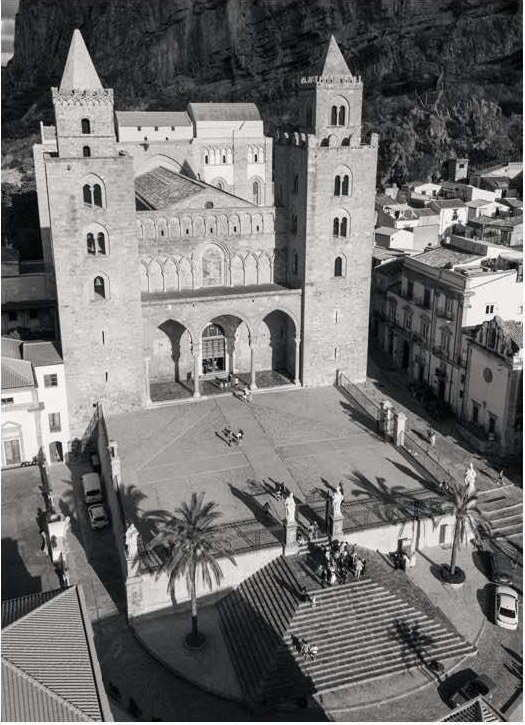Black and white view of the twin Norman towers and arched facade of a historic Sicilian cathedral above a palm-dotted piazza