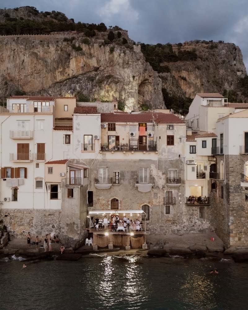 Seafront stone houses at dusk beneath a towering rocky headland with a lit terrace wedding reception over the water in Sicily