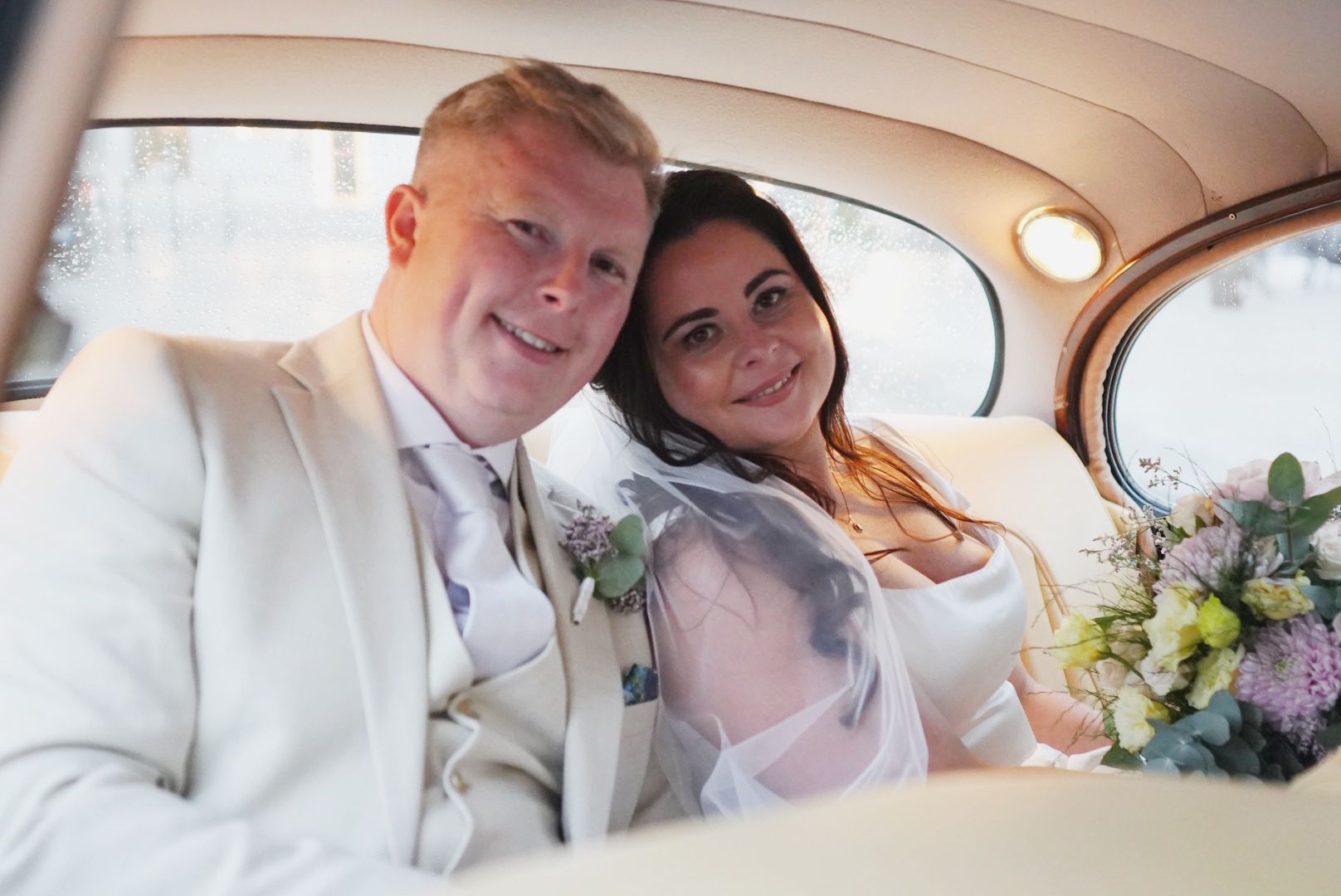 Gráinne and Phil together inside a cream vintage wedding car, Gráinne holding a pastel bouquet, on their way from the ceremony