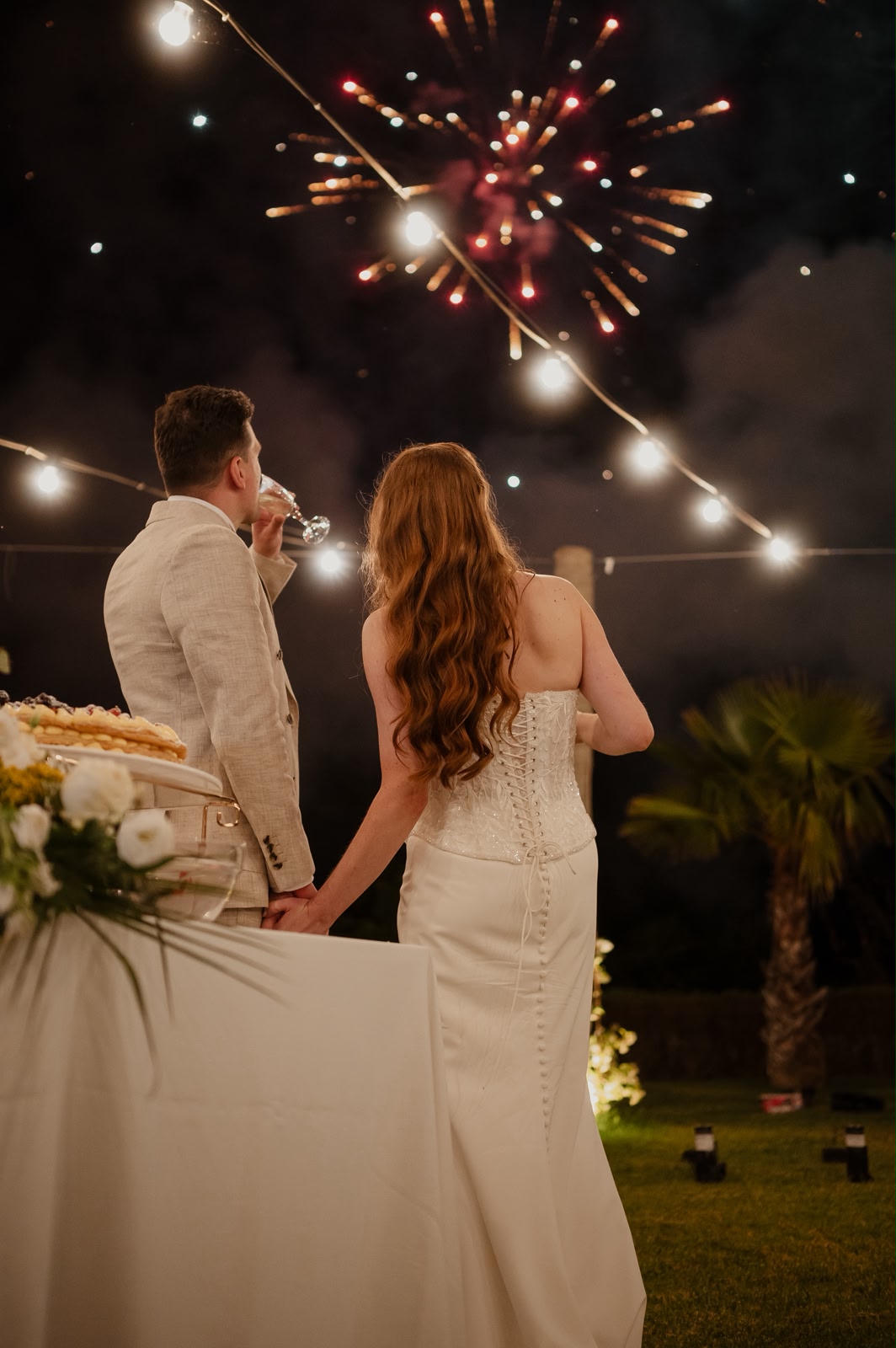 Jessica e Seb di spalle mentre guardano i fuochi d’artificio illuminare il cielo notturno