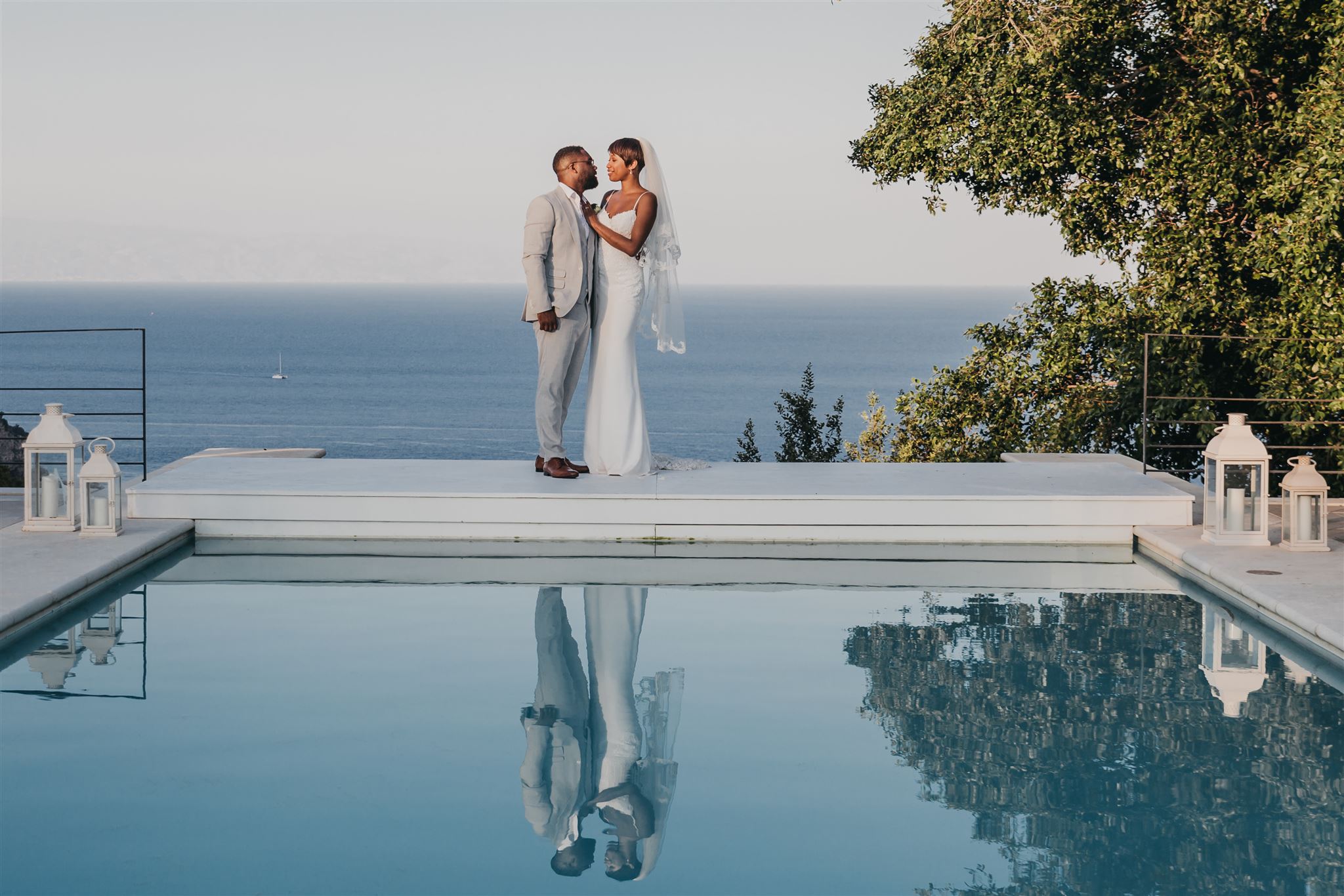 Kysha and Shem standing together at the edge of a reflecting pool at Villa Mon Repos, the calm Ionian Sea stretching to the horizon behind them