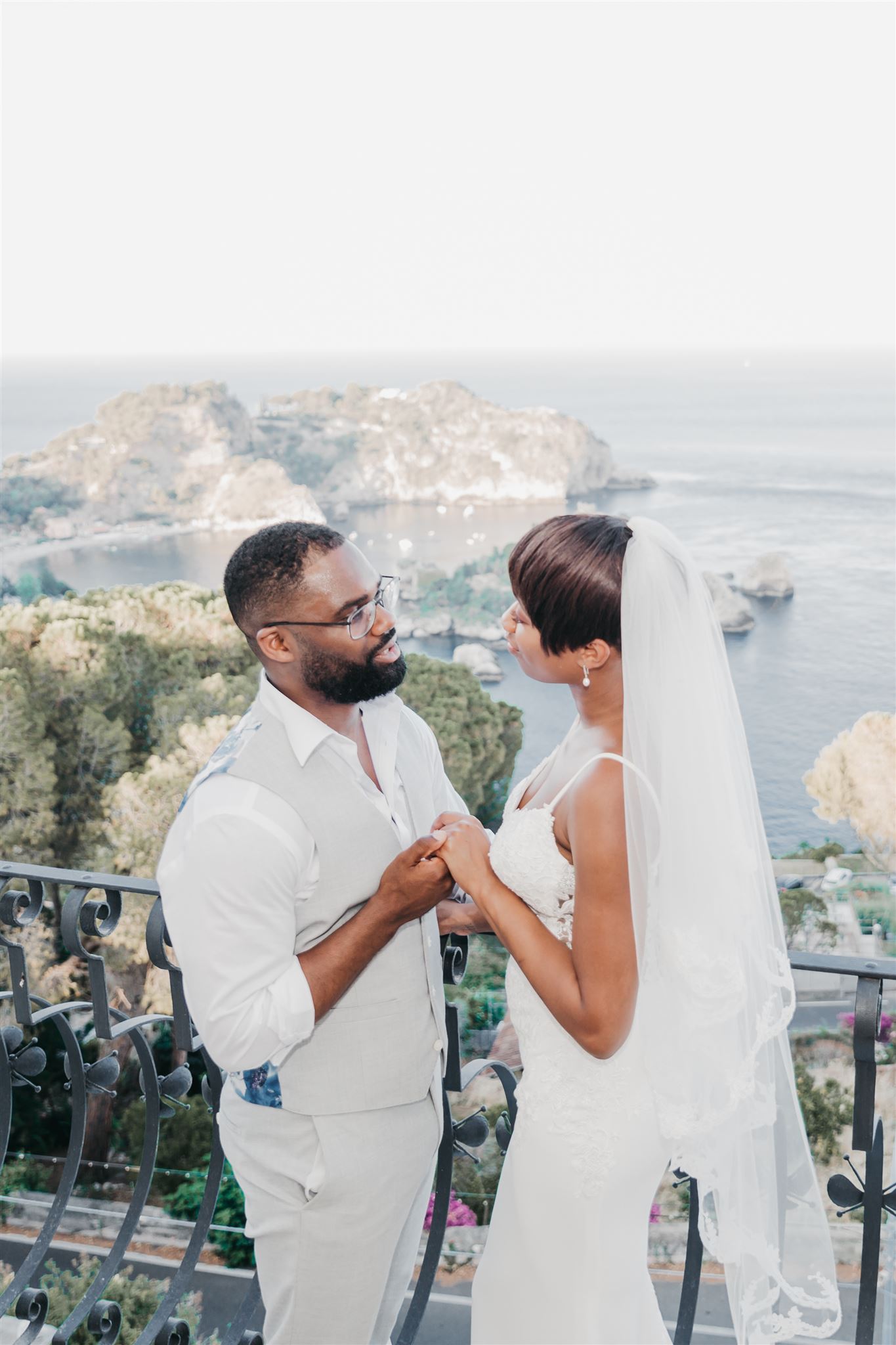 Kysha and Shem holding hands on a wrought-iron villa balcony in Taormina, with Isola Bella and the bay visible in the background