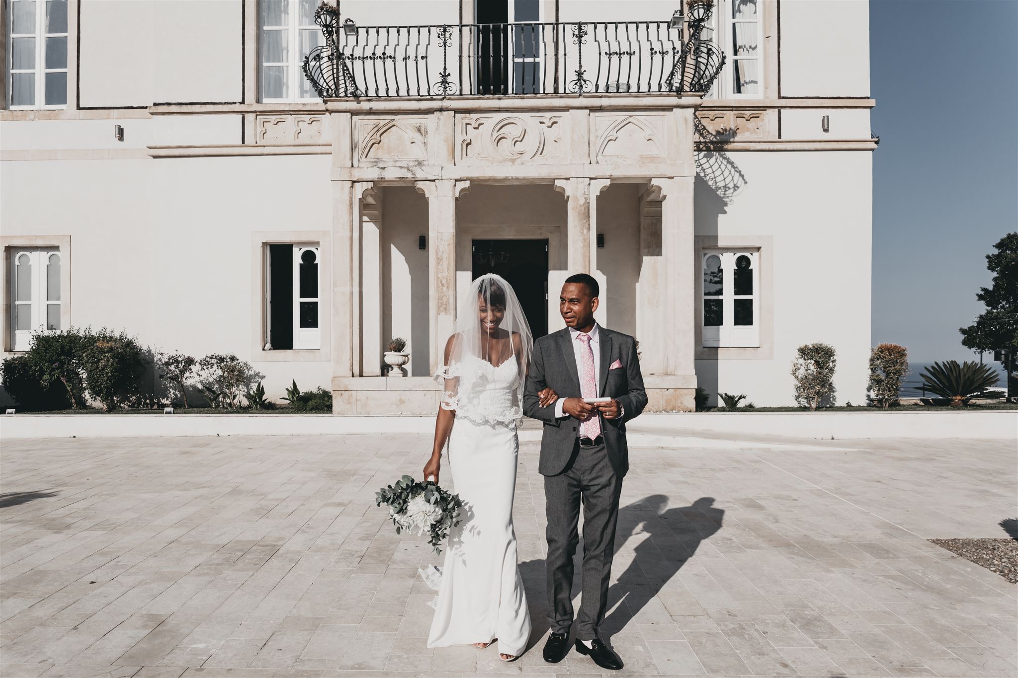 Kysha in her lace gown and veil walking arm in arm across the sunlit terrace in front of the elegant white facade of Villa Mon Repos