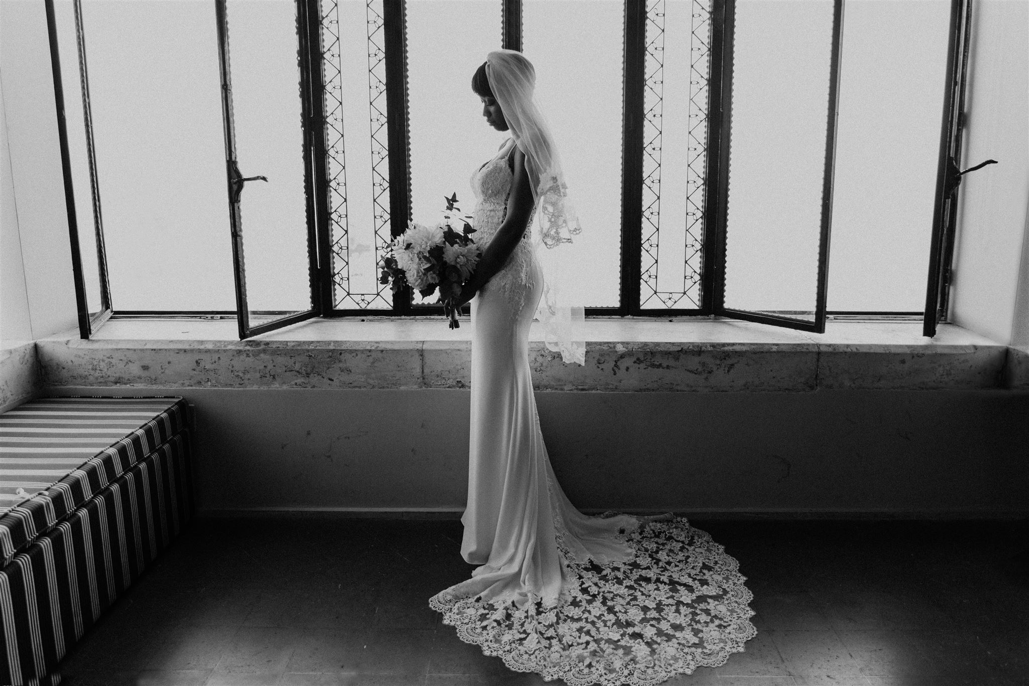 Elegant black and white portrait of a bride in a fitted lace gown and cathedral veil standing beside a tall arched window
