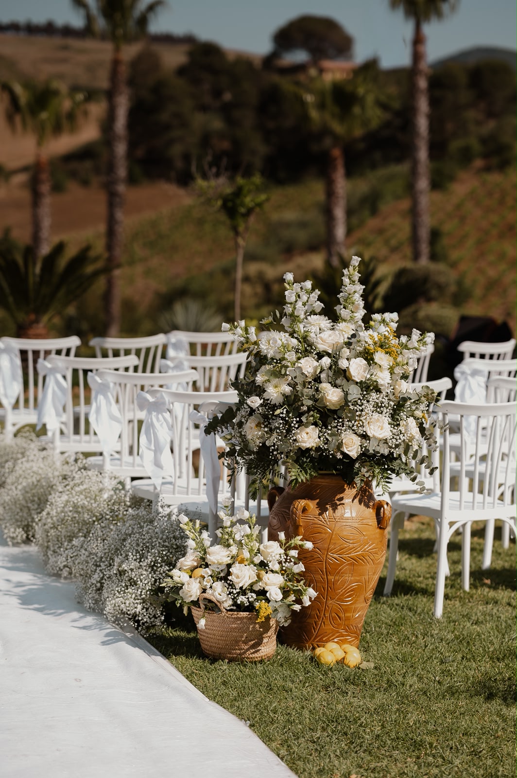 Elegant white ceremony florals in a rustic terracotta urn with Sicilian lemons lining the aisle of white chairs
