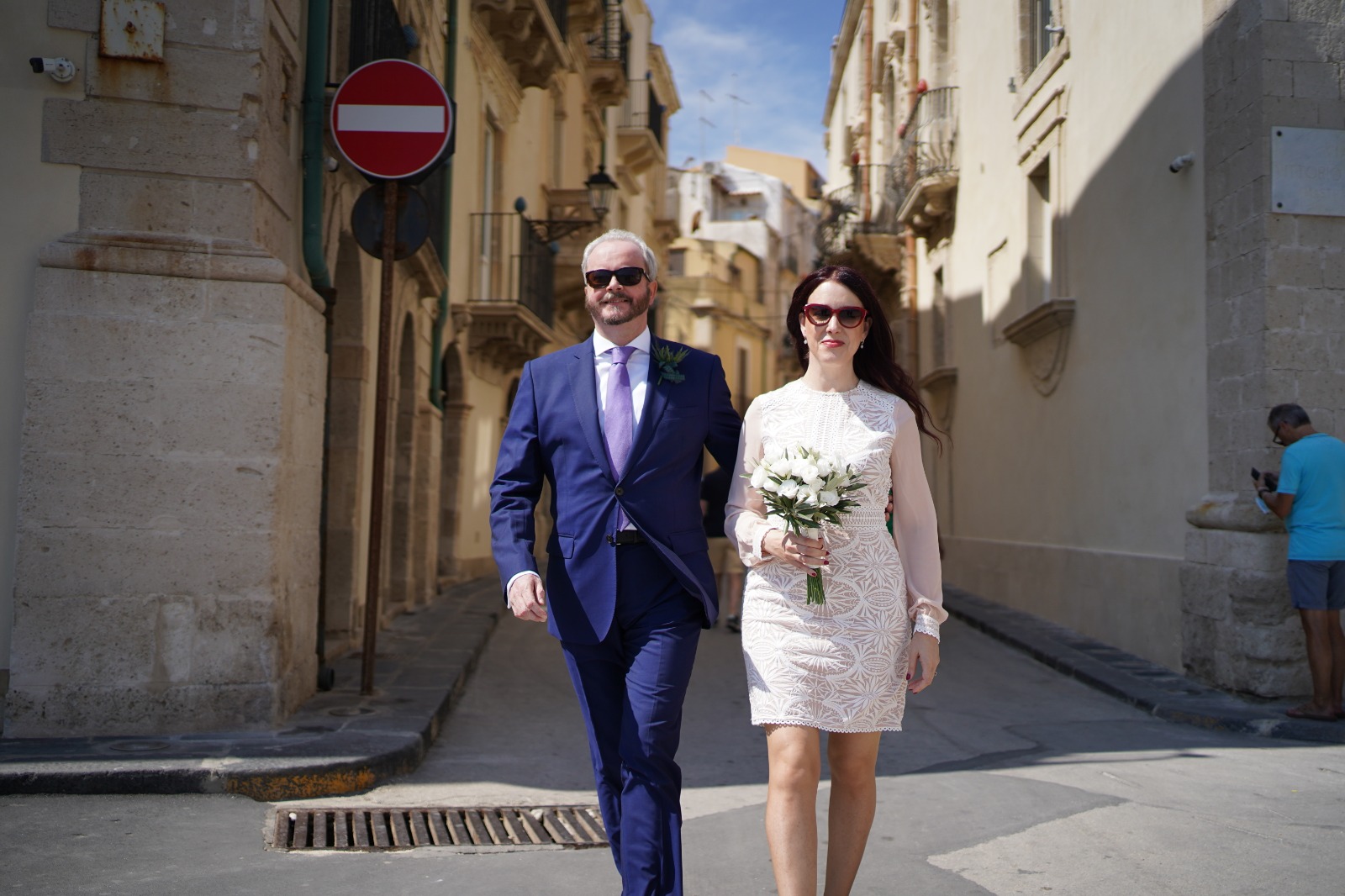 Stylish newlyweds walking hand in hand through a sunlit historic Sicilian street, the groom in a navy suit and lilac tie, the bride in a white lace dress