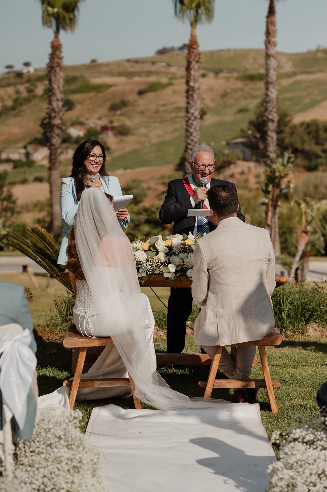 Sabina officiating an outdoor wedding ceremony with a microphone alongside an Italian official, in the Sicilian hills