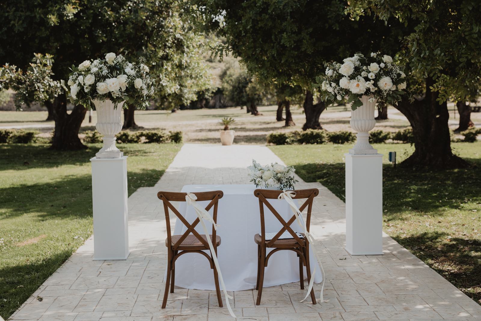 Tavolo per la cerimonia nuziale sotto gli alberi nei giardini del Borgo del Carato, vicino a Siracusa, con fiori bianchi su piedistalli in pietra