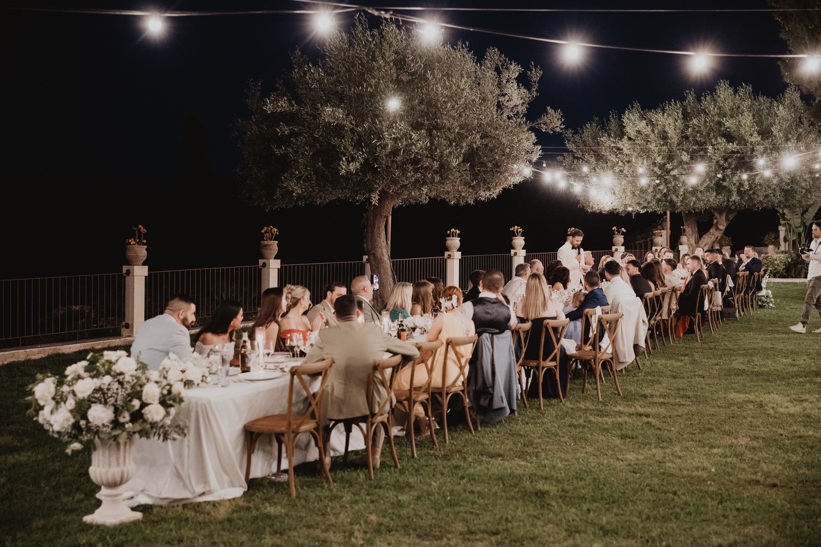 Ospiti a cena sotto le luci sospese a una lunga tavolata del Borgo del Carato, dopo il tramonto