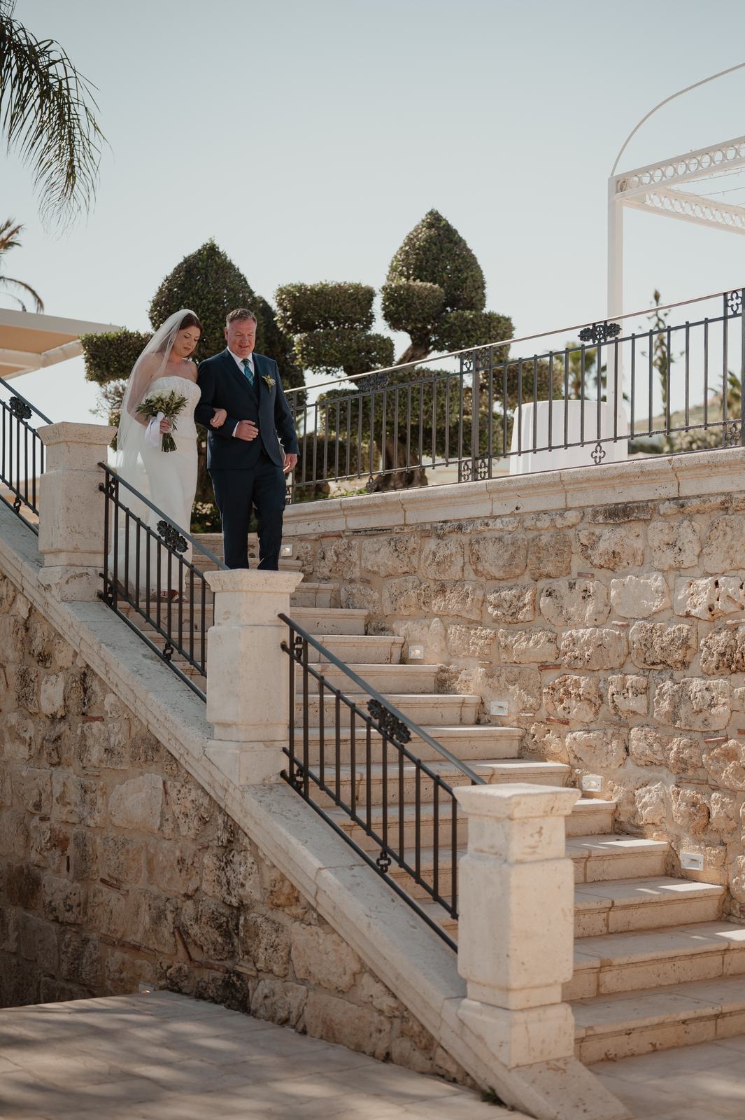 La sposa scende la scalinata in pietra al braccio del padre per il rito civile al Casale degli Aranci, nella Sicilia occidentale